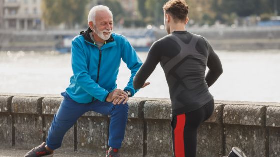 Un homme d'une soixantaine d'années s'étire en extérieur, accompagné par une personne plus jeune qui guide le mouvement.