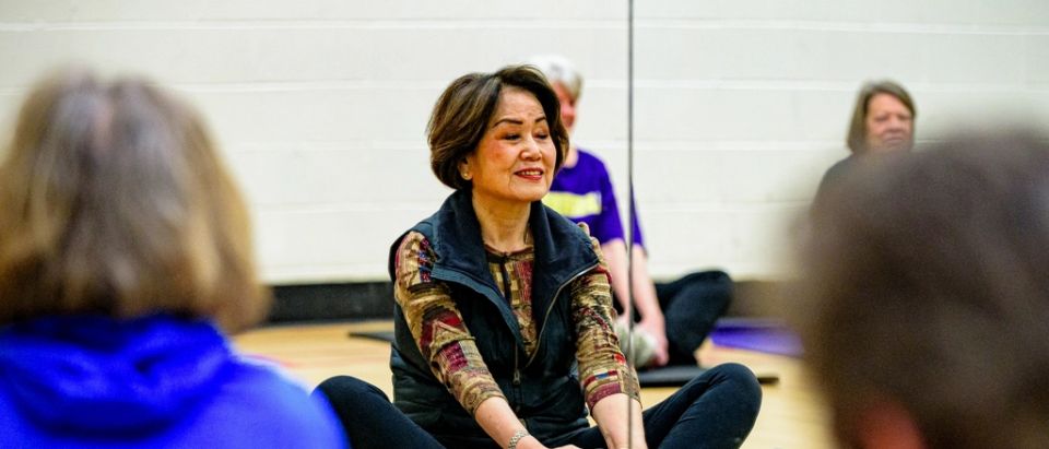 Une femme étire ses jambes, assise sur un tapis, pendant un cours collectif en salle.